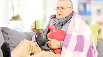 Winter - man sitting inside with a blanket, scarf, hot water bottle, and hot drink
