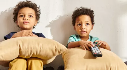Two young children watching TV, sitting on a couch with pillows and the TV remote