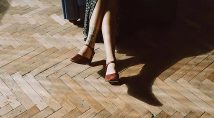 Sun shines on a woman with tattoos on her legs, as she sits on a herringbone wood floor. March 2024