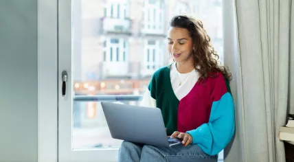 A woman working on a laptop while sitting on a windowsill