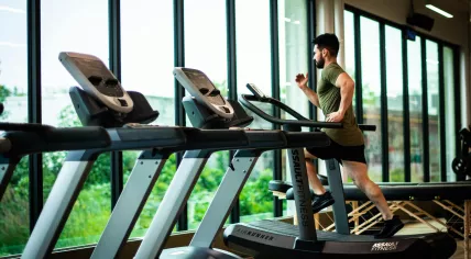 A man running on a curved treadmill inside a fitness center, with a massage table and weight bench