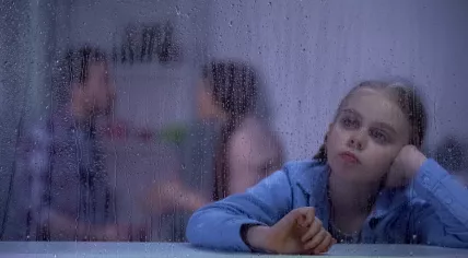 A young girl looks through a rain-covered window as her parents argue in the background.