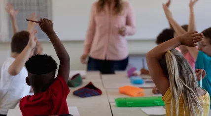 Back view of children in a primary school classroom raising their hands to answer the teacher's question