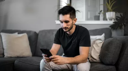 A concerned man sitting on a couch staring at a mobile phone. Jan. 2023