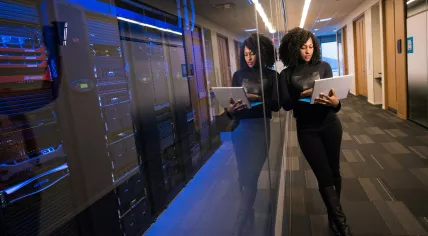 A woman using a Microsoft Surface laptop while standing in front of an office server and tech room. Nov. 2019
