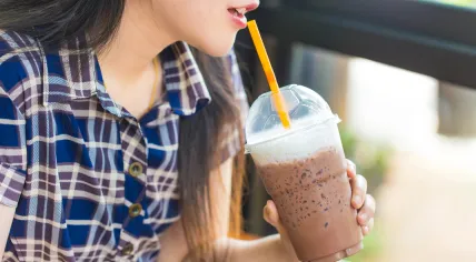 A girl drinks an iced coffee from a plastic cup using a plastic straw.