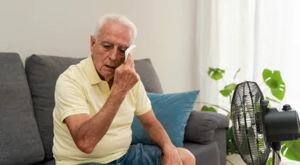 An elderly man wiping sweat from face while sitting on sofa near an electric fan at home