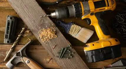 A battery-powered electric drill and a hammer on a workbench with other tools. 2022