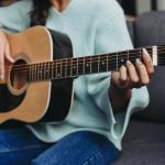 Cropped image of a young woman in turquoise sweater playing acoustic guitar on sofa 