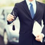 Car rental concept - man in a suit with a car key and clipboard standing in front of a car