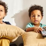 Two young children watching TV, sitting on a couch with pillows and the TV remote