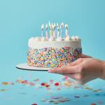 Partial view of woman holding birthday cake with candles on blue background with confetti