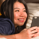 A smiling young woman on a bed holding her phone