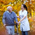 A caregiver walks with an elderly man through a forest.