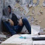 A spoon, lighter, syringe, and a bag of white powder lie in the foreground, while a man sits in the background, partially out of focus.