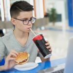 Teenage boy eating a burger and drinking a soft drink while sitting with a laptop in a food court