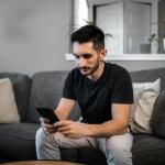 A concerned man sitting on a couch staring at a mobile phone. Jan. 2023