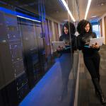 A woman using a Microsoft Surface laptop while standing in front of an office server and tech room. Nov. 2019