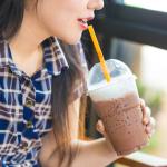 A girl drinks an iced coffee from a plastic cup using a plastic straw.