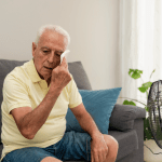 An elderly man wiping sweat from face while sitting on sofa near an electric fan at home