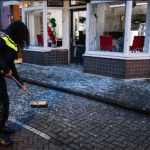 A police officer sweeping up glass from a window shattered in an explosive attack