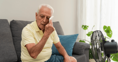 An elderly man wiping sweat from face while sitting on sofa near an electric fan at home